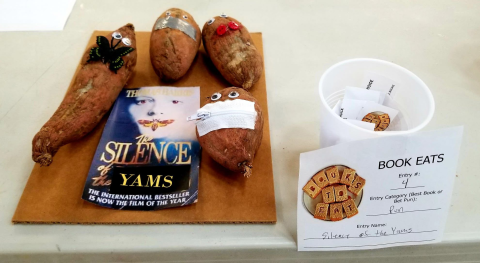 Sweet potatoes with faces and mouths covered lay on a board with a cover of the Book Silence of the Lambs. A white card next to the potatoes reads Silence of the Yams
